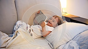 Portrait of happy laughing toddler boy lying in bed with digital tablet