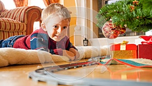 Happy laughing toddler boy lying on floor and looking on toy railroad around big Christmas tree