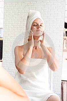 Happy laughing young woman in white bath towels doing morning make up applying face cream looking at the mirror