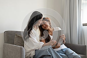 Happy lesbian couple hugging having fun using digital tablet relaxing on couch at home. Two smiling asian women friends