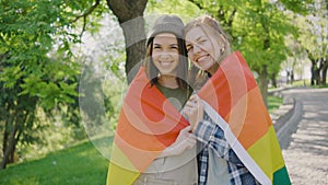 Happy lesbian couple hugging and holding rainbow flag in park and looking at the camera at sunrise