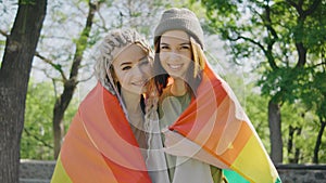 Happy lesbian couple hugging and holding rainbow flag in park and looking at the camera at sunrise