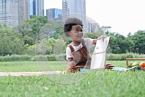 Happy little African kid boy holding a brush to paint in book while sitting on picnic mat in park