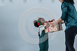 Happy little African kid girl with green bib clothes and earmuffs get gift box from Asian mother in Christmas holiday
