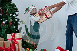 Happy little African kid girl with green bib clothes and reindeer Santa hat get gift box from Asian senior father in Xmas holiday