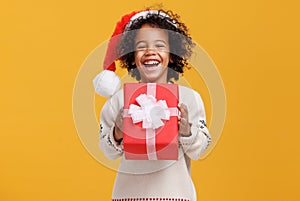 Happy little afro american curly boy in santa hat and knitted sweater hugging big Christmas gift box tightly