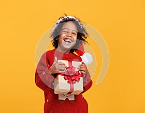 Happy little afro american curly boy in santa hat and knitted sweater hugging big Christmas gift box tightly