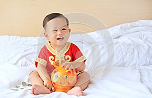 Happy little Asian baby boy in traditional Chinese dress with a piggy bank sitting on bed at home. Kid saving money concept