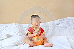 Happy little Asian baby boy in traditional Chinese dress putting some coins into a piggy bank sitting on bed at home. Kid saving
