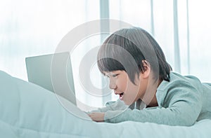 Little Asian boy is using laptop computer on his bed in his room in daytime by the windows