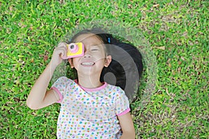 Happy little Asian child girl with digital camera lying on green lawn background