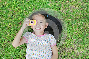 Happy little Asian child girl with digital camera lying on green lawn background