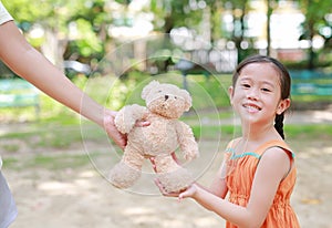 Happy little Asian child girl get a teddy bear doll from her mother in the park outdoor. Surprise gift from mom for daughter