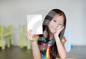 Happy little Asian child girl holding blank white paper card in her hand. Kid showing empty paper note copy space in children room