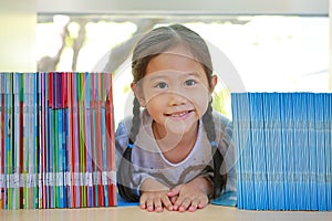 Happy little Asian child girl lying on bookshelf at library. Children creativity and imagination concept