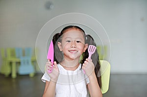 Happy little Asian child girl showing plastic knife and spade of frying pan in play room ready to cook learning