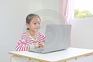 Happy little asian child girl sitting at desk and using laptop computer stay at home