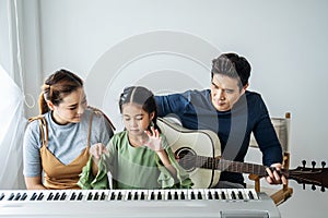 Happy little Asian daughter playing piano with mother and father play guitar at home, Mother teaching daughter to play piano,