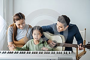 Happy little Asian daughter playing piano with mother and father play guitar at home, Mother teaching daughter to play piano,