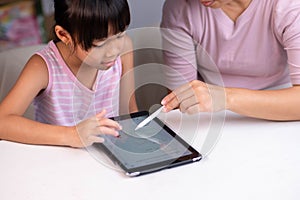 Happy little Asian daughter talking with mother using digital tablet to do homework or study online. Happy young girl and adult