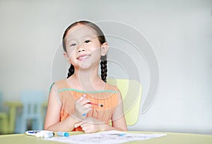 Happy little Asian girl drawing and painting with water color on paper in children room with looking at camera