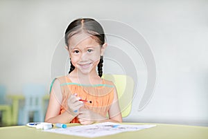 Happy little Asian girl drawing and painting with water color on paper in children room with looking at camera