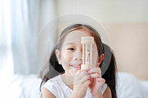 Happy little Asian girl holding sandglass in hand with looking through camera. Waiting times with hourglass