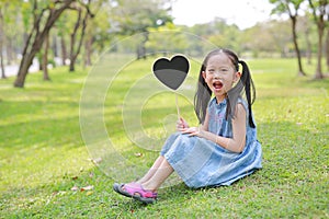 Happy little Asian kid girl holding blank heart label sitting on green grass at garden outdoor