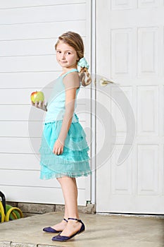 Happy little beautiful girl holds apple and stands white door