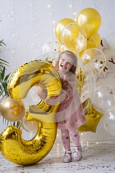 happy little blond girl in pink dress holding big gold foil balloon number and digit three 3