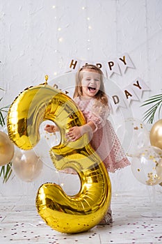 happy little blond girl in pink dress holding big gold foil balloon number and digit three 3