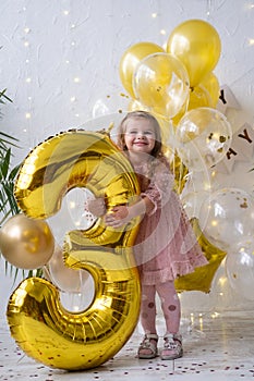 happy little blond girl in pink dress holding big gold foil balloon number and digit three 3