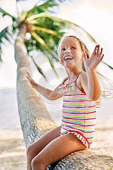 Happy little blonde girl playing on the beach sitting on palm tree.
