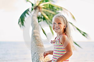 Happy little blonde girl playing on the beach sitting on palm tree.