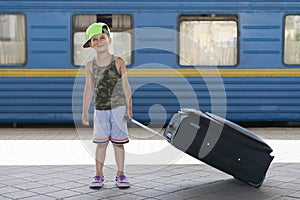 Happy little boy with a big black suitcase on a background of a blue train. Adventure and Travel Concept