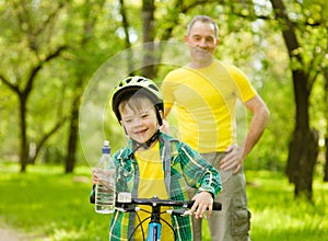 Happy little boy with a bottle of water is learning to ride a bike with his grandfather