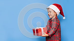 A happy little boy in Christmas pajamas and a hat with a gift box. A place for your text. Studio shot isolated on a blue