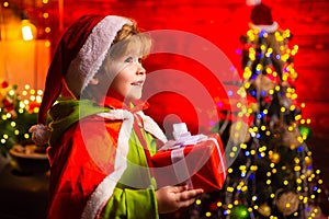 Happy little boy by the Christmas tree with his Christmas gift, curios. Little kid is wearing Santa clothes. Christmas