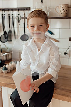 Happy little boy congratulating smiling mother and giving card with red heart during holiday celebration at home