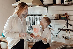 Happy little boy congratulating smiling mother and giving card with red heart during holiday celebration at home