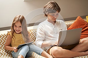 Happy little boy and girl, brother and sister sitting on sofa at home, using laptop and digital tablet and watching