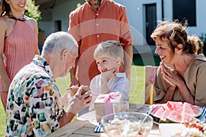 Happy little boy giving birthday present to his senior grandfather at generation family birthday party in summer garden