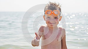 Happy little boy in glasses for swimming shows gesture - thumb up on the background of the sea. Travel and summer vacation concept