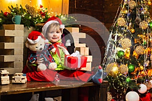 Happy little boy with his Teddy bear and Christmas gift is looking at the camera. Happy cute child in Santa hat with