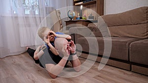 A happy little boy lies on his dad's back and hugs him at home on the floor.