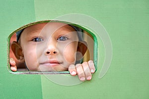 Happy little boy looks through window in green wooden house