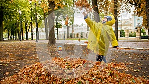 Happy little boy in raincoat playng and throwing autumn leaves at park