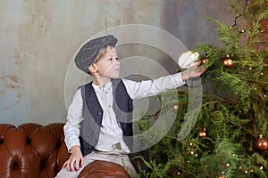 Happy Little boy sit on sofa and decorates Christmas tree in Cozy living room. Smiling Little blond boy in white shirt with vest a