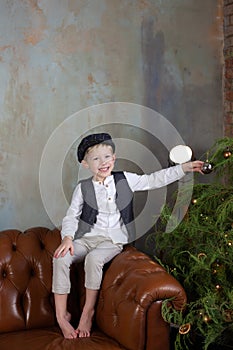 Happy Little boy sit on sofa and decorates Christmas tree in Cozy living room. Smiling Little blond boy in white shirt with vest a