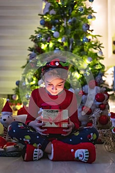 Happy little boy sitting in front of a christmas tree and suprisingly looking at the gift